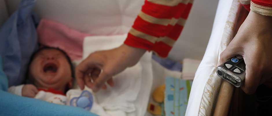 Majka se naginje nad uplakanom bebom držeći u ruci pumpu za kontrolu inzulina | Tending the Crying Baby with the Insulin Pump Controller in Hand